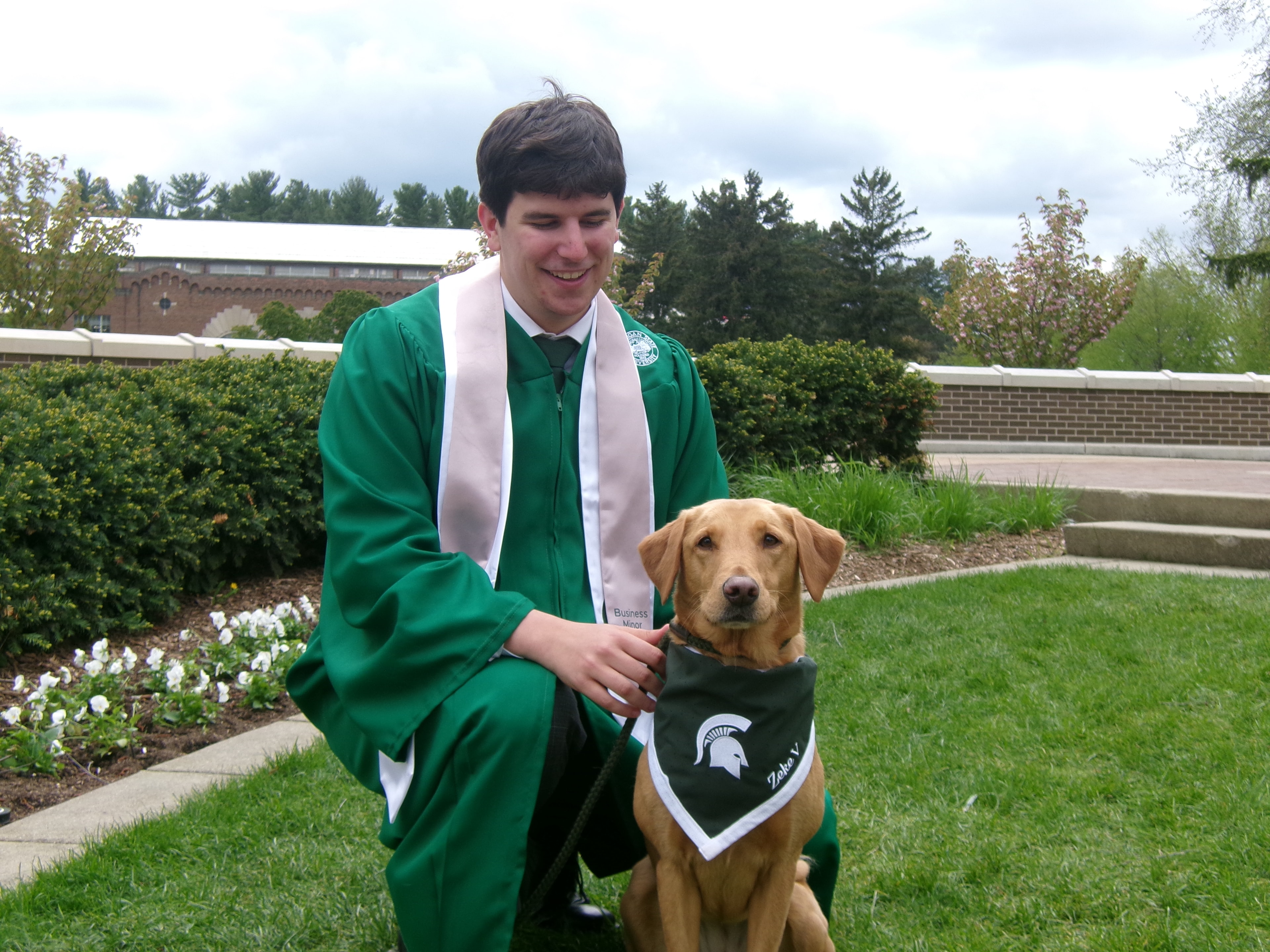 MSU graduate in cap and gown posing with Zeke the WonderDog on campus